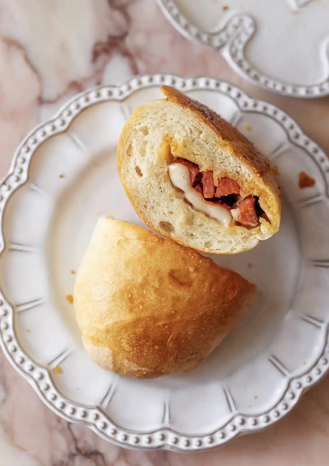 A golden-brown bread roll, sliced in half and placed on a white decorative plate, reveals melted cheese and chunks of meat…