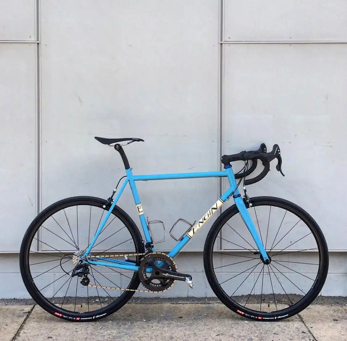 A light blue road bicycle with black components and gold chain is leaning against a plain white wall. The brand name…