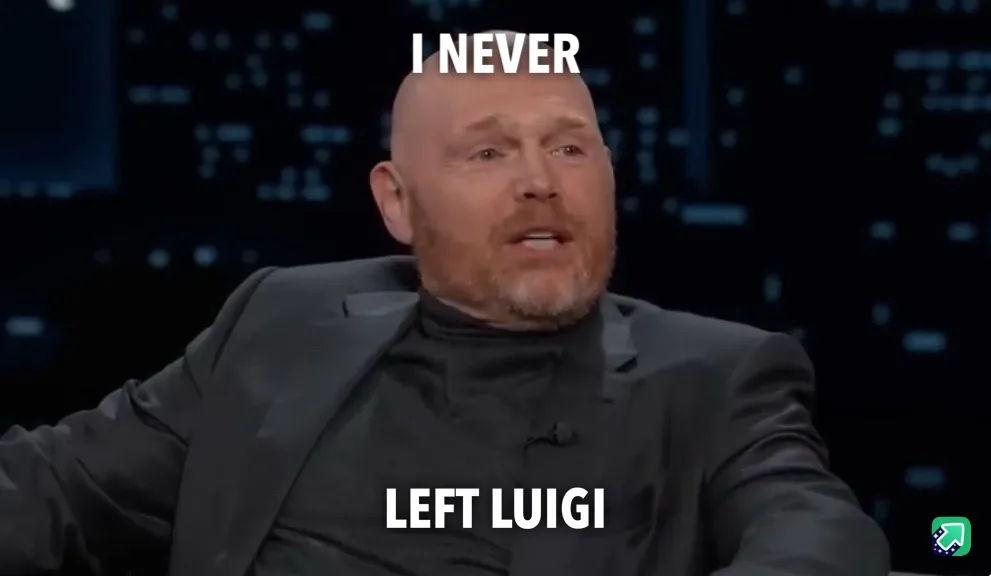 A bald man with a beard wearing a black suit and turtleneck sits in a chair against a dark background with blue lights…