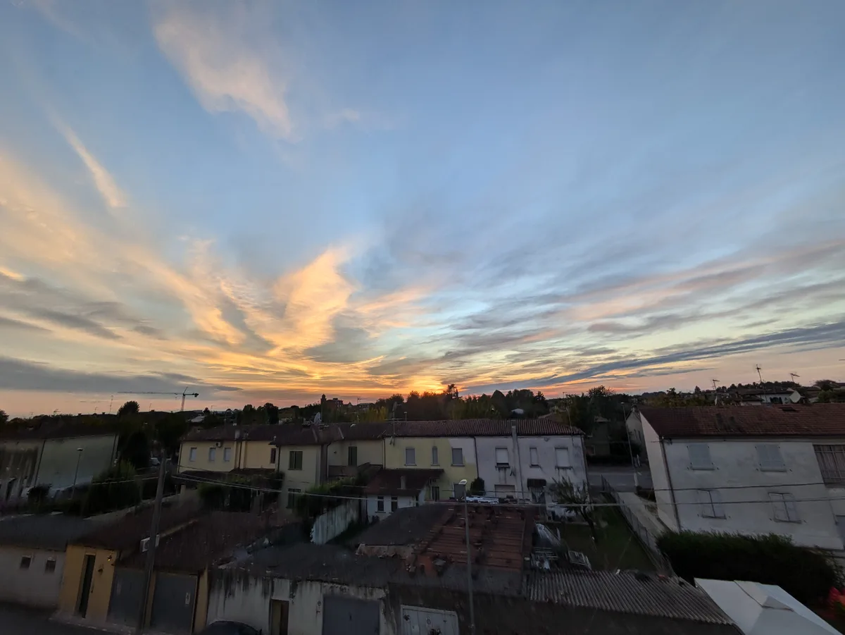 A photograph of a residential neighborhood at sunset, with colorful orange and blue clouds above houses with tiled roofs…