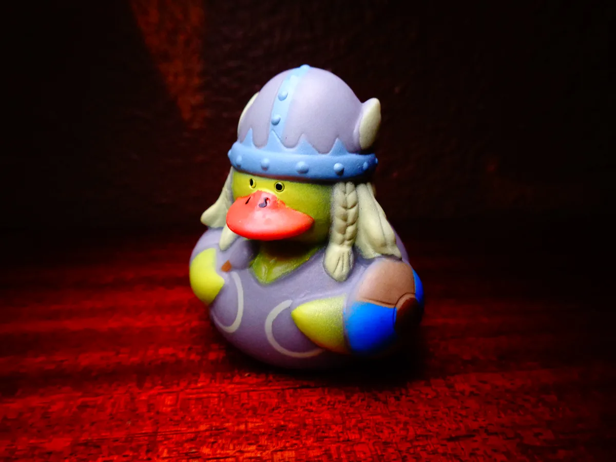 A green rubber duck is dressed as a viking, wearing a purple helmet with horns and braided blonde hair, standing on a red…