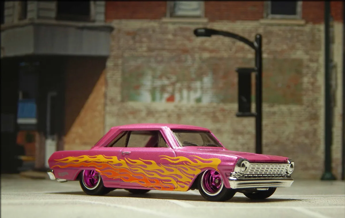 a pink hot wheels toy car with orange and yellow flame decals is shown parked on a miniature street in front of a brick…