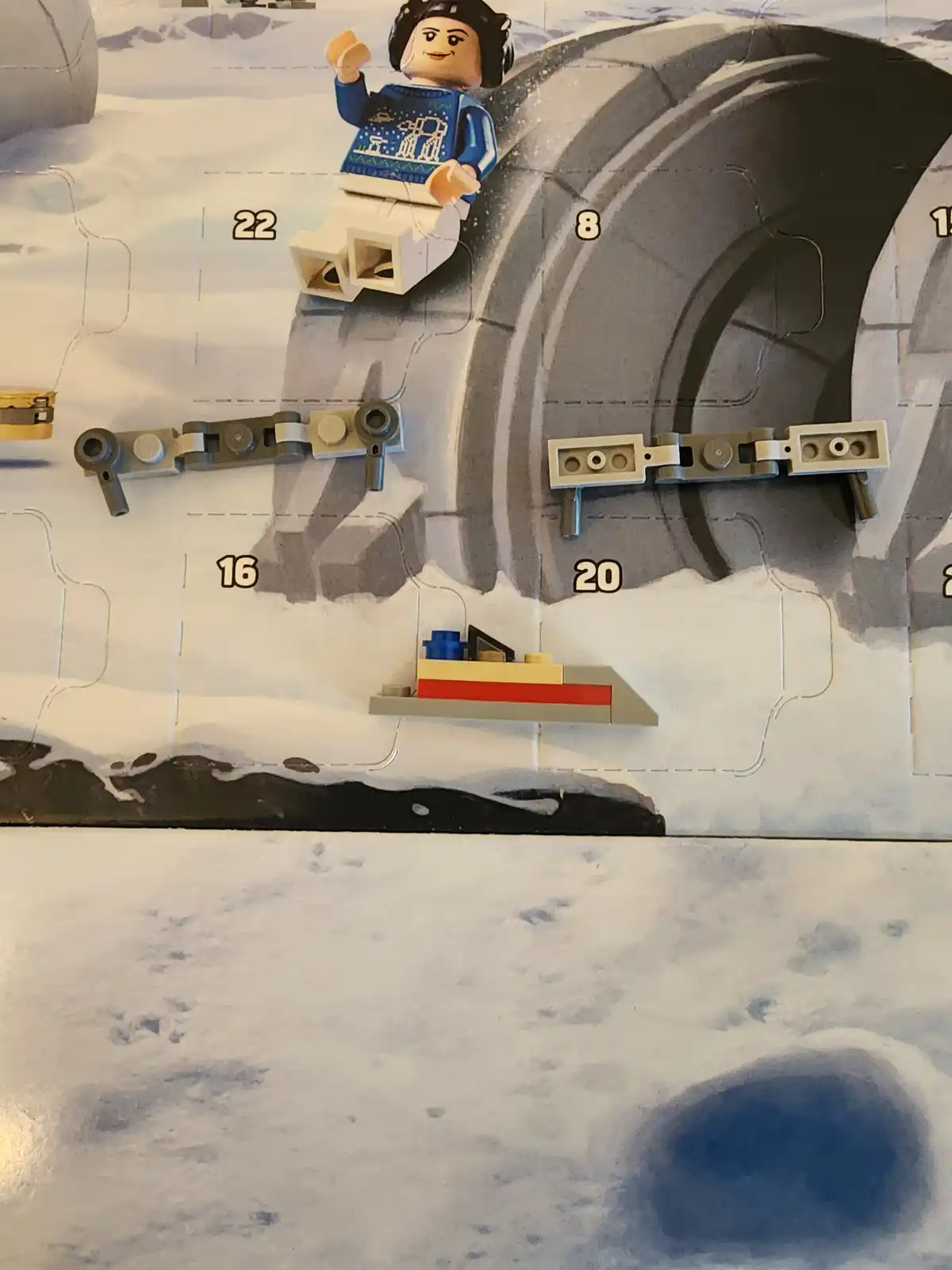 A close-up of a LEGO Star Wars Advent Calendar showing a minifigure in a blue sweater with a snowflake pattern, sitting on a…