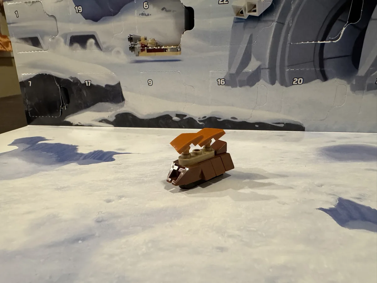 A small, brown and orange LEGO spaceship model sits on a white, snowy surface in front of a partially opened LEGO Advent…