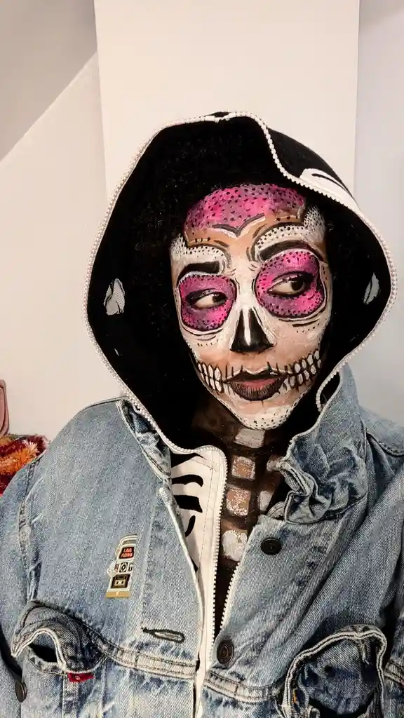 A person with dark curly hair wears a black bucket hat and a blue denim jacket over a white shirt with a skeleton print…