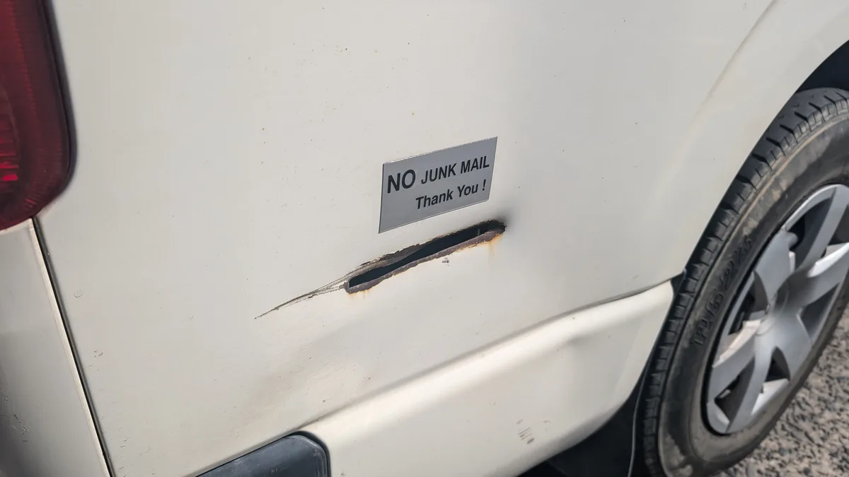 A close-up of a white vehicle's side panel shows a rectangular metal sign reading 'NO JUNK MAIL Thank You !' directly above…