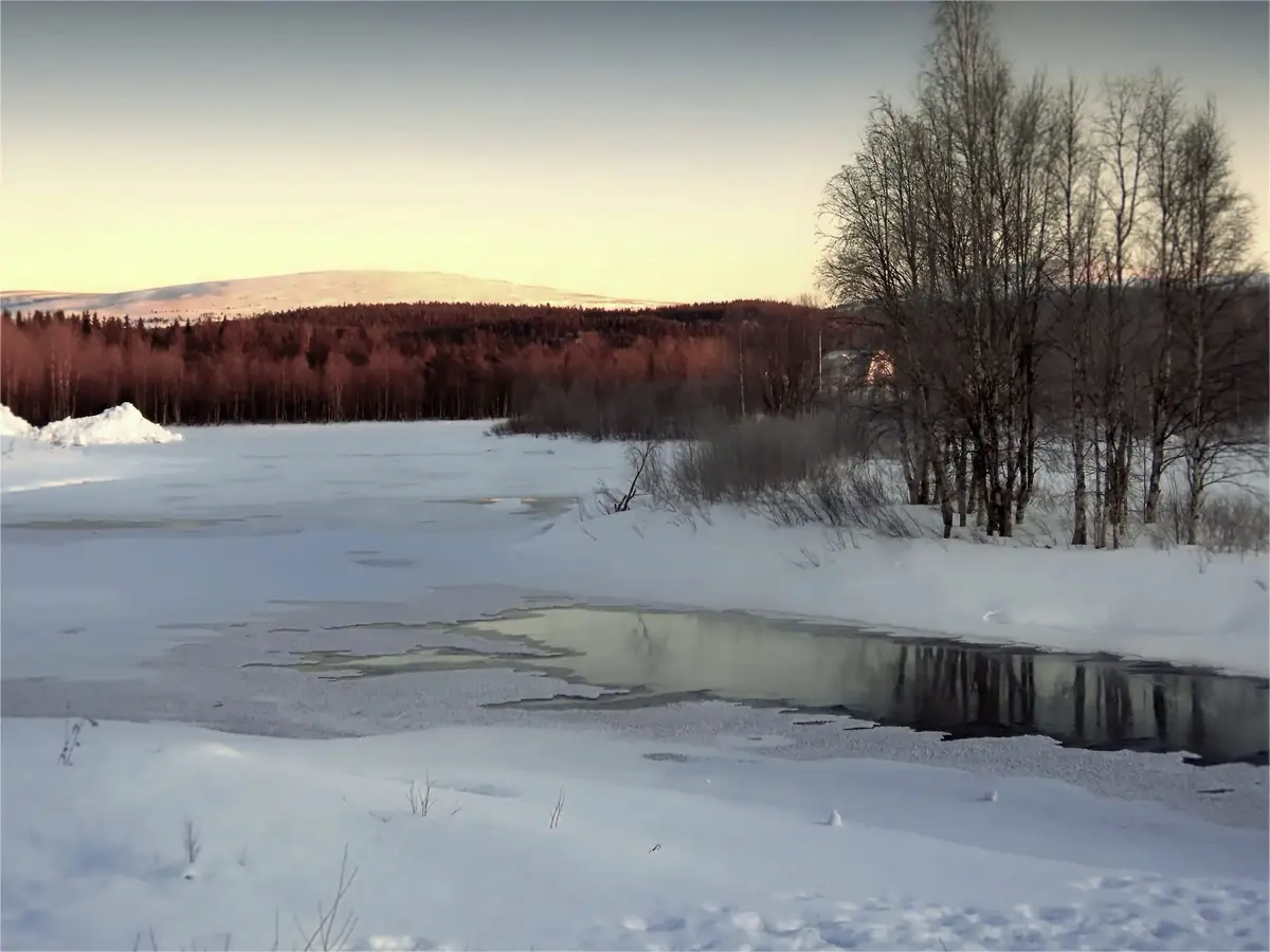 A serene winter landscape shows a snow-covered frozen lake with patches of open water reflecting bare trees, distant hills…