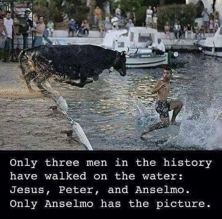 A black bull leaps from a sandy shore into water, splashing dramatically. A shirtless man runs through the water nearby…