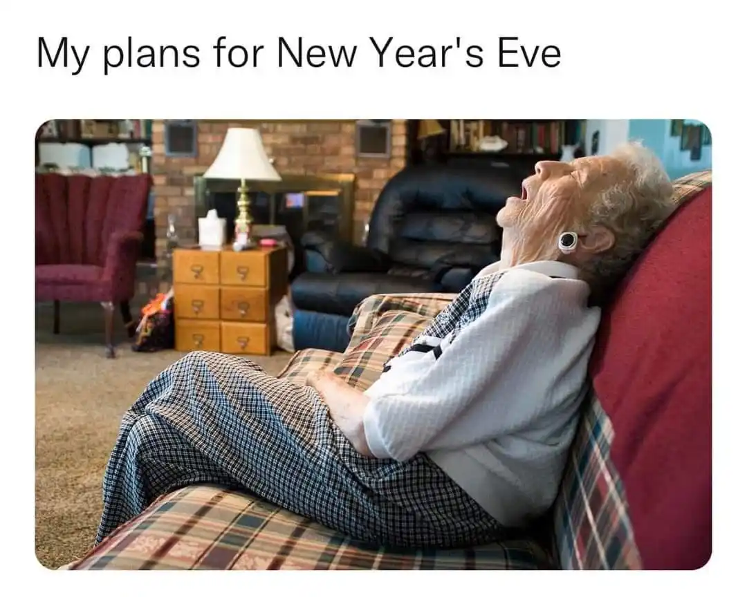 An elderly woman with white hair and a white sweater is lying back on a red couch, mouth wide open in a yawn or laugh…