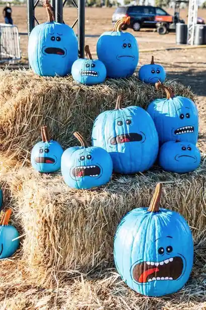 Several blue pumpkins, painted to resemble the character Blue from Adventure Time, are arranged on hay bales outdoors. Each…