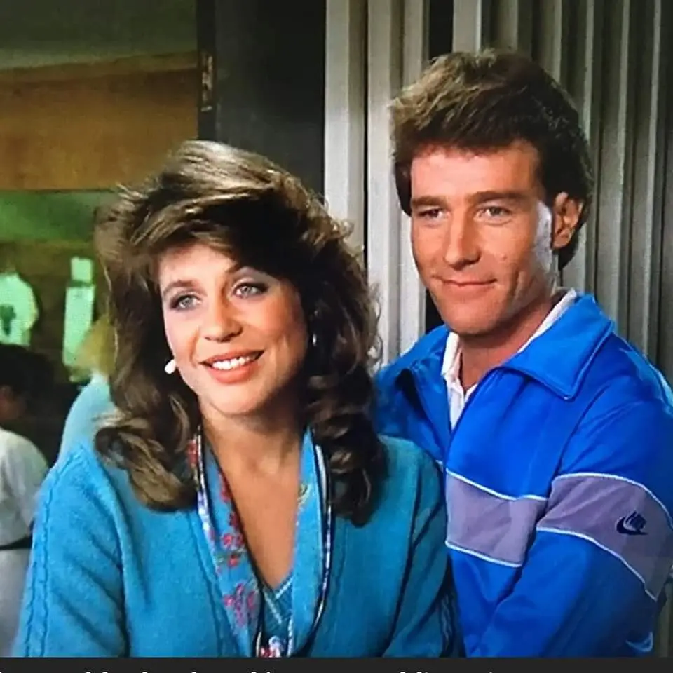 A still from a 1980s TV show featuring a woman with voluminous brown hair wearing a blue cardigan and a man with styled hair…