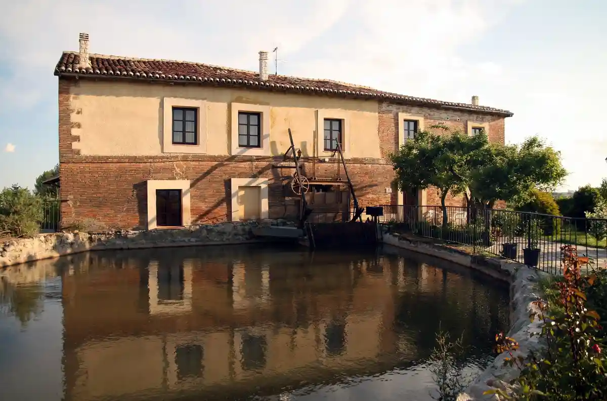 A serene photo of a two-story brick building with a tiled roof, situated beside a calm canal with a waterwheel attached to…