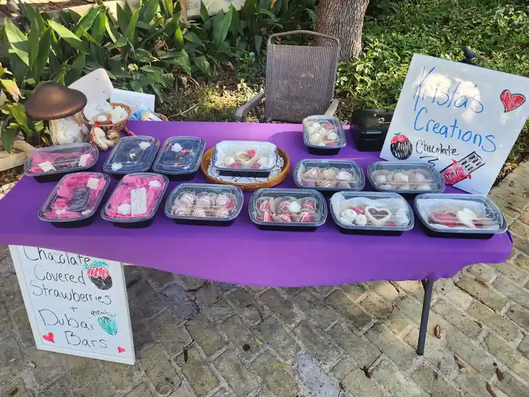 An outdoor table covered in a purple cloth displays numerous clear plastic containers filled with assorted chocolate-covered…
