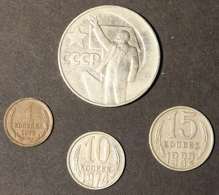 Four Soviet-era coins are arranged on a dark surface: a large 1975 1 kopeck coin, a 1974 10 kopeck coin, a 1982 15 kopeck…