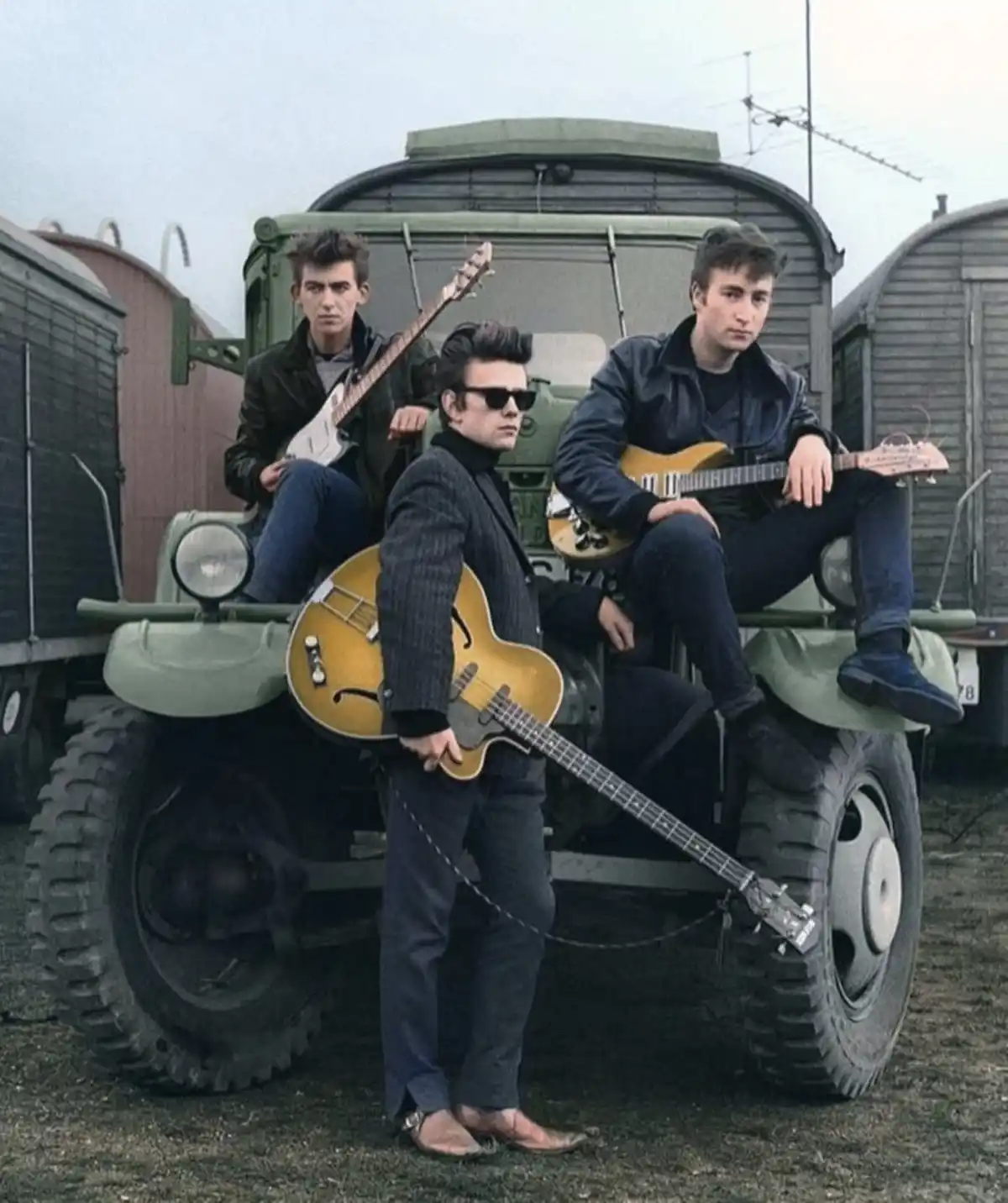 Three young men, identified as The Beatles, pose with guitars on and around a green military-style truck in a yard with…