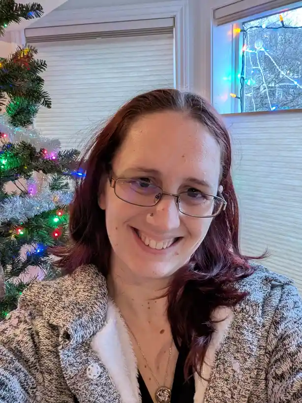 A woman with reddish-brown hair and glasses smiles at the camera while standing in front of a decorated Christmas tree with…
