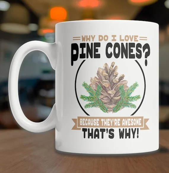 A white ceramic mug with a handle sits on a wooden surface, featuring a printed design with a pine cone and pine needles…