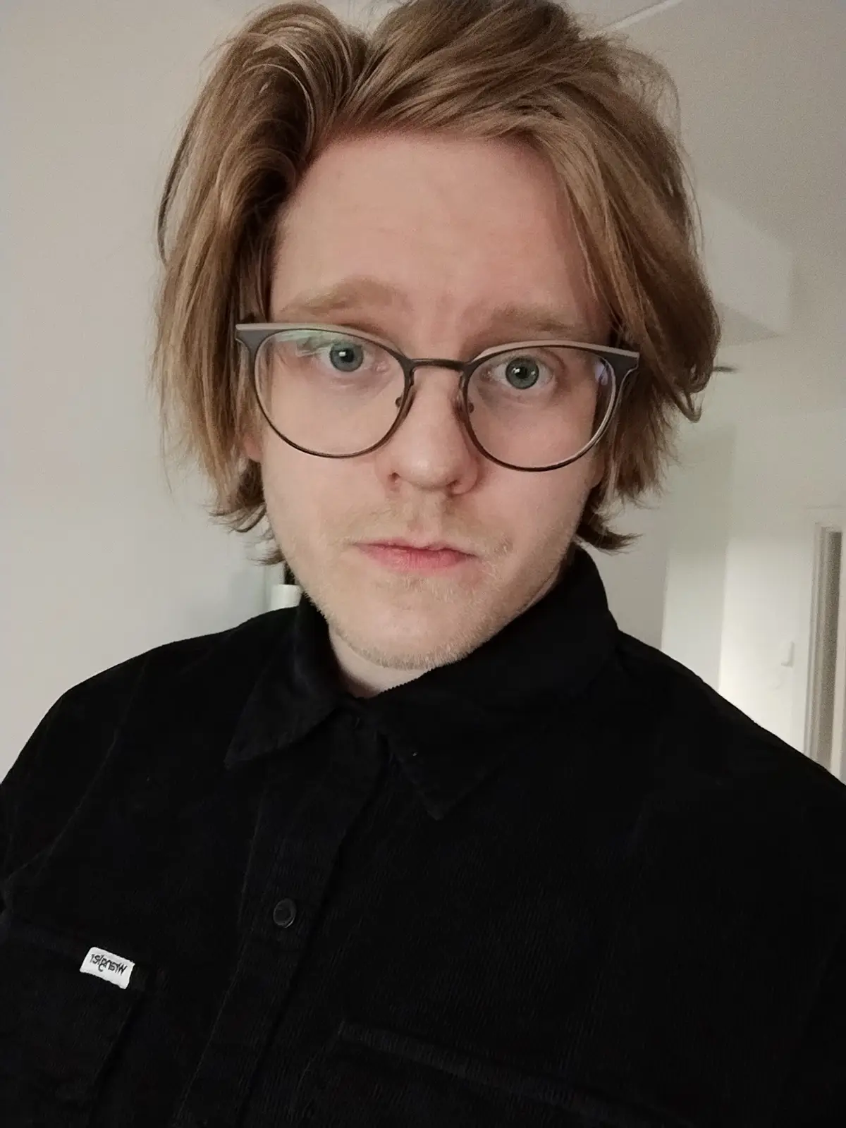 A close-up selfie of a young man with light brown hair, wearing round-framed glasses and a black corduroy shirt, staring…
