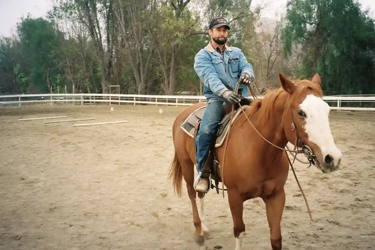 A man with a beard and baseball cap rides a brown horse with a white blaze in a sandy riding arena surrounded by trees and…