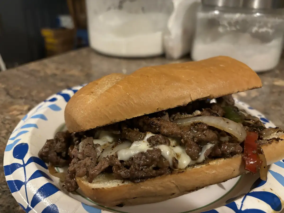 A close-up photo of a Philly cheesesteak sandwich on a paper plate, filled with sautéed meat, melted cheese, onions, and…
