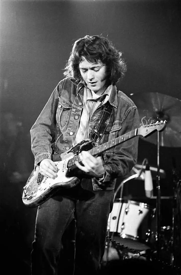 A black-and-white photo of a man with long hair wearing a denim jacket, playing an electric guitar on stage with a drum kit…