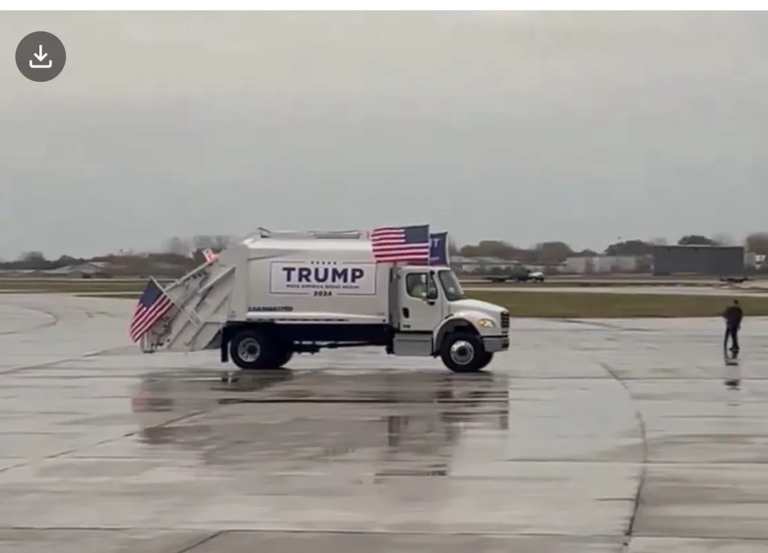 A white garbage truck is driving on a wet tarmac, decorated with American flags and a large sign reading 'TRUMP MAKE AMERICA…