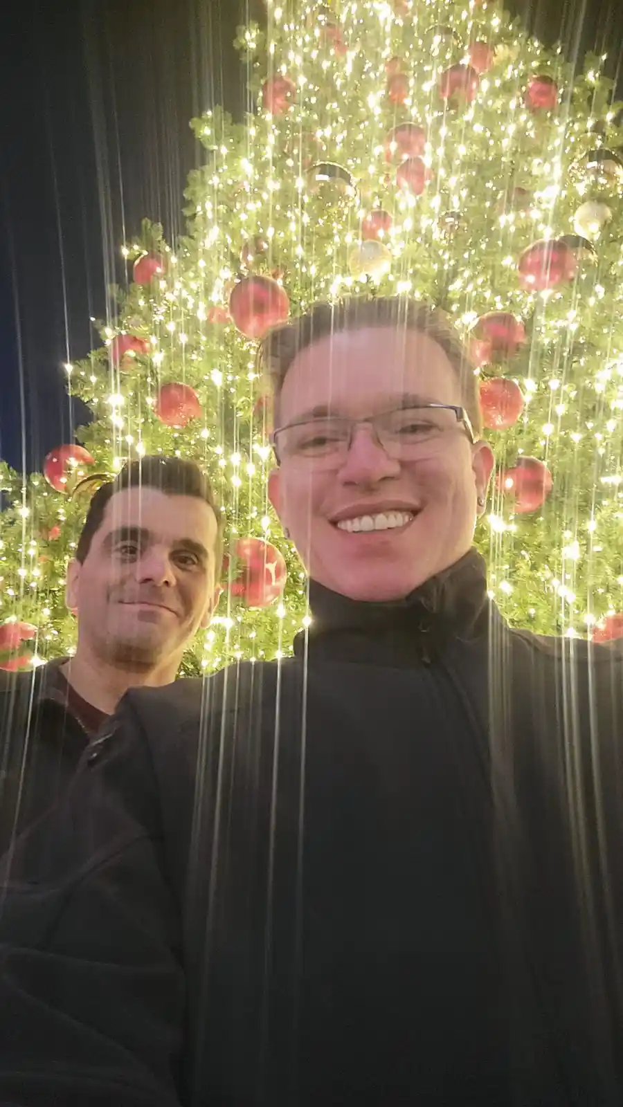 Two men pose for a selfie in front of a brightly lit, decorated Christmas tree. Vertical streaks of light, resembling rain…
