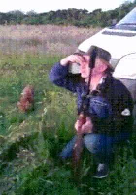A man in a hat and jacket crouches in a grassy field beside a white vehicle, holding a rifle. In the background, a small…