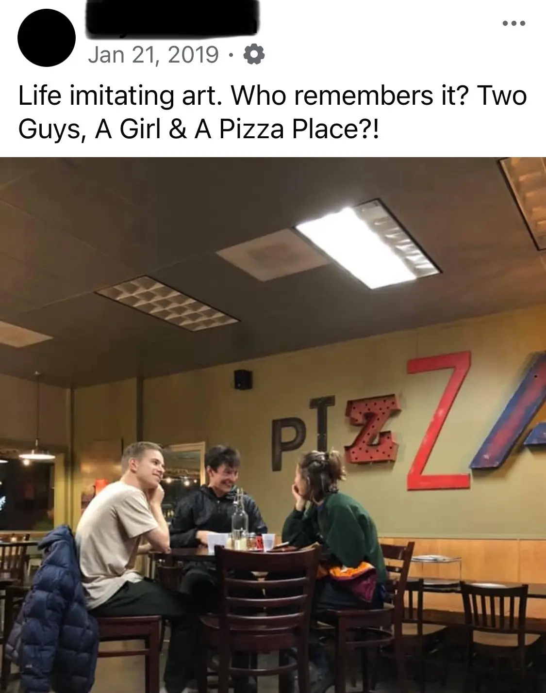 Three young people sit at a table in a pizza restaurant, smiling and chatting. Behind them, a large wall sign spells out…