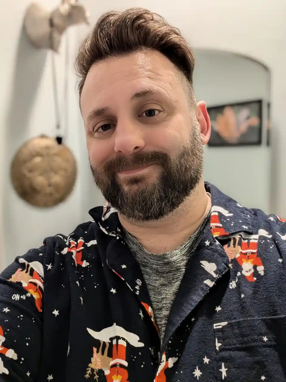 A man with a beard and styled hair smiles at the camera while wearing festive pajamas featuring Santa Claus and reindeer…