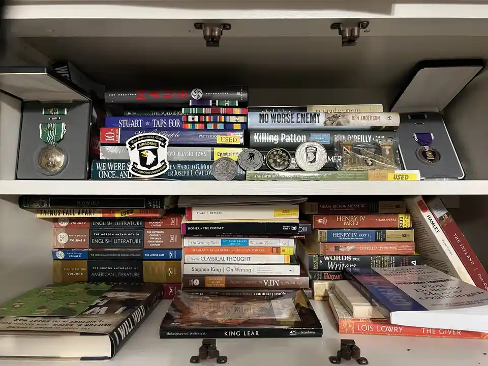 A white bookshelf is filled with books on literature, history, and military topics, interspersed with military medals and…