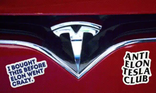 A close-up of a red Tesla car's front grille showing the silver Tesla logo. Two white stickers are overlaid: one on the left…