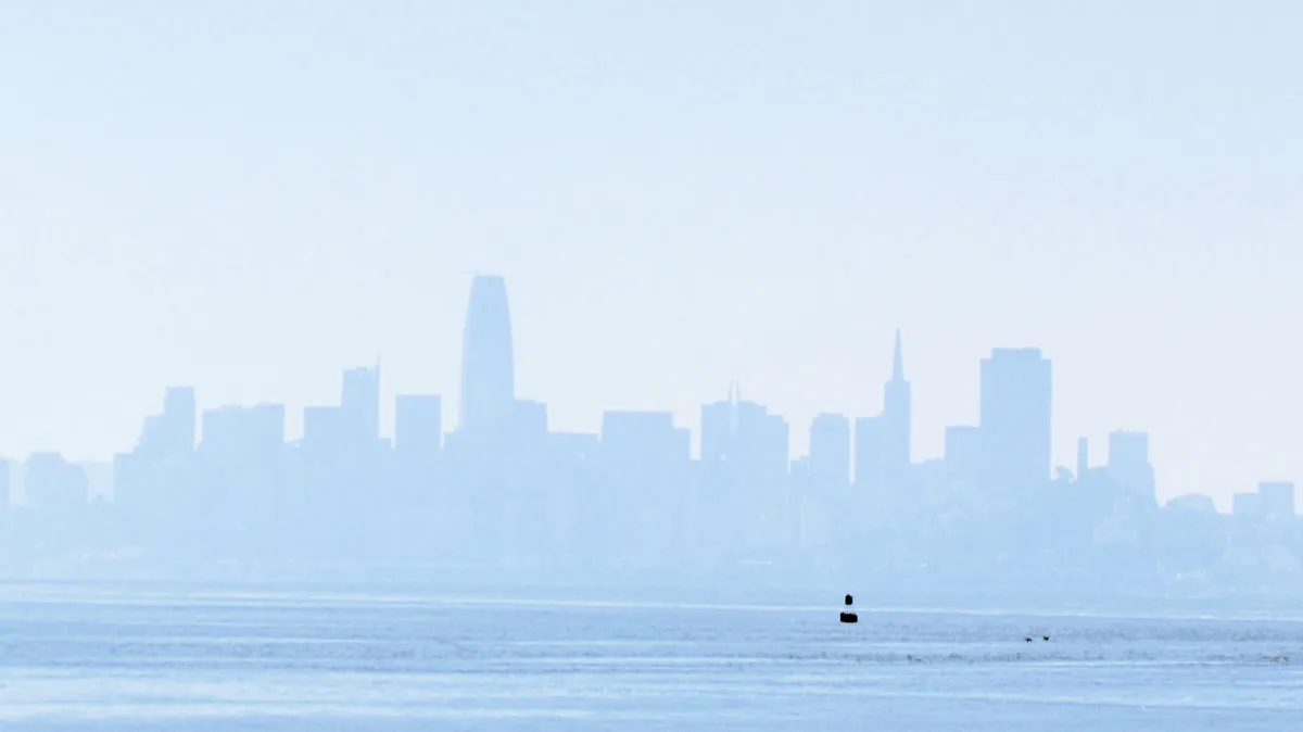 A hazy, blue-tinted photo shows a distant city skyline with recognizable skyscrapers like the Salesforce Tower and…