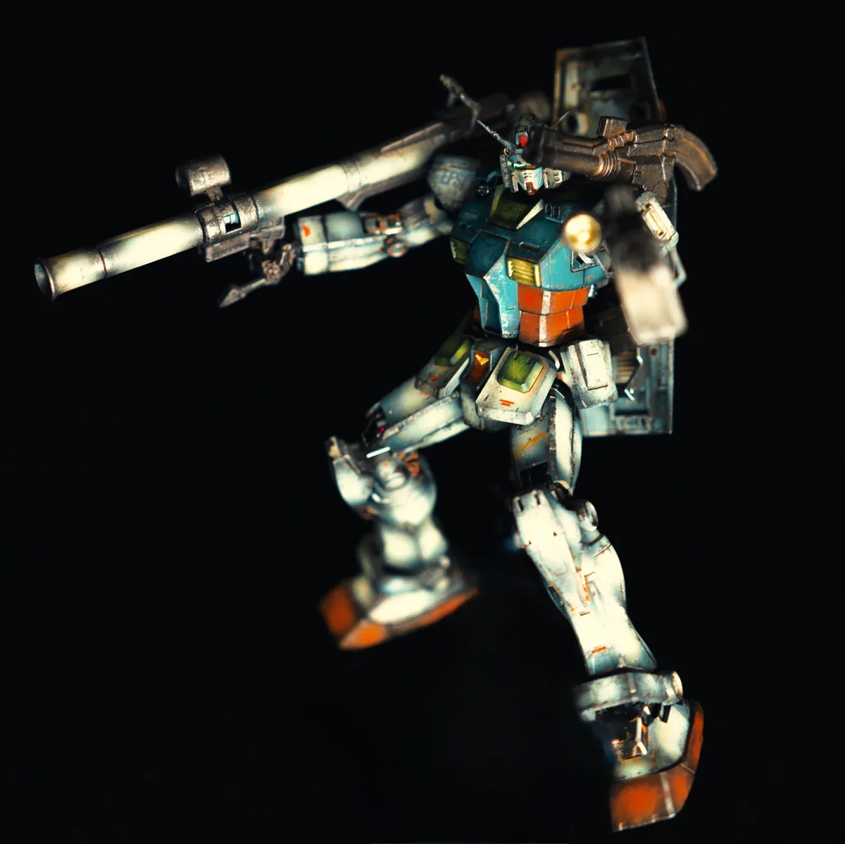 A weathered, blue and orange Gundam model stands against a black background, holding a large rifle. The text overlay reads…