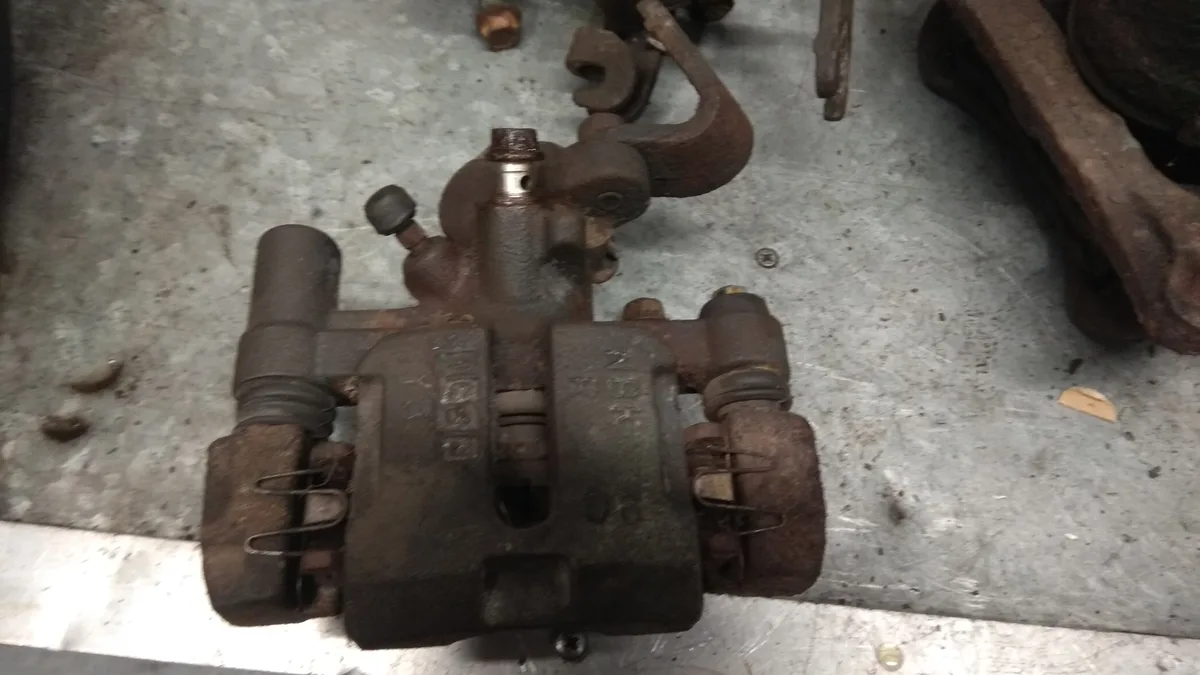 A heavily rusted, dirty automotive brake caliper lies on a scratched metal workbench, surrounded by other rusty mechanical…