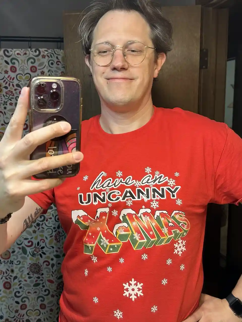 A man with glasses takes a mirror selfie wearing a red t-shirt that reads 'have an UNCANNY X-MAS' with snowflake graphics…