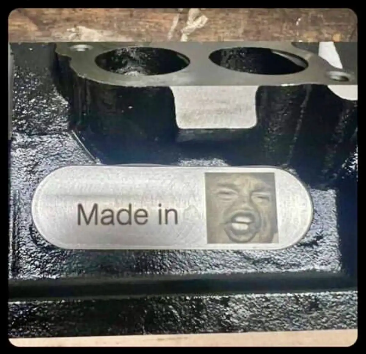 A close-up photo of a black metal engine part with a silver oval label. The label reads 'Made in' followed by a small square…