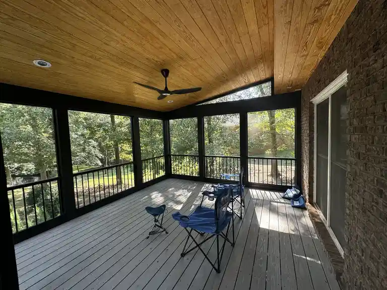 An empty screened porch with wooden flooring, black-framed windows, and a wooden ceiling with a ceiling fan. Two blue…