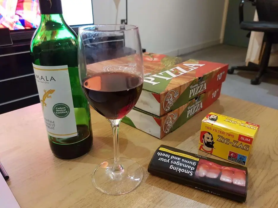 A wooden table holds a green wine bottle, a glass of red wine, two stacked pizza boxes, and a pack of Zig-Zag slim cigarette…