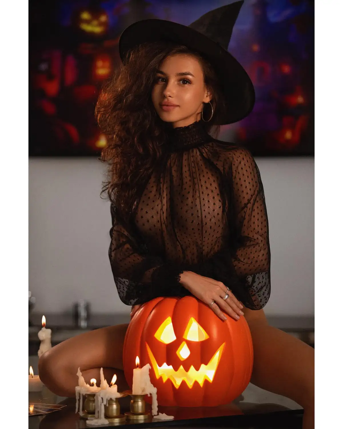 A woman with long dark hair wearing a black witch's hat and a sheer black polka-dot top is posing with one hand resting on a…