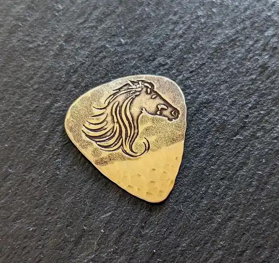 A close-up photo of a gold-colored guitar pick with an engraved horse head design, resting on a dark gray slate surface. The…
