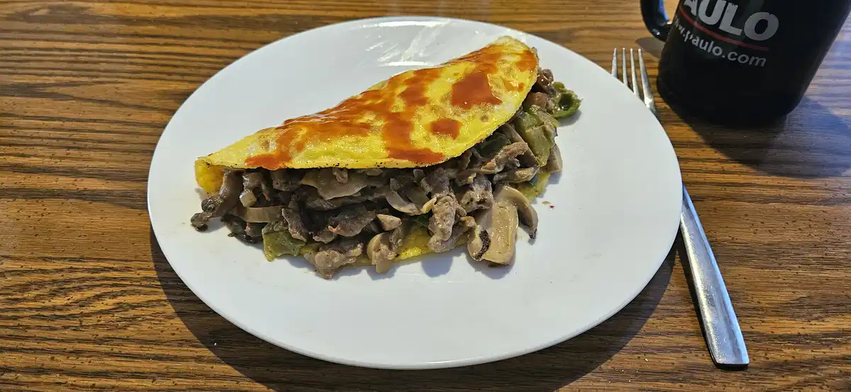 A close-up photo shows a large, folded omelette on a white plate, filled with meat, mushrooms, and green peppers, drizzled…