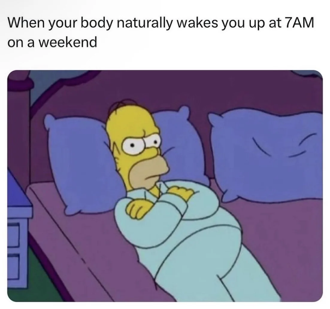 Homer Simpson lies in bed looking annoyed and groggy, wearing light blue pajamas, with purple pillows behind him. The…