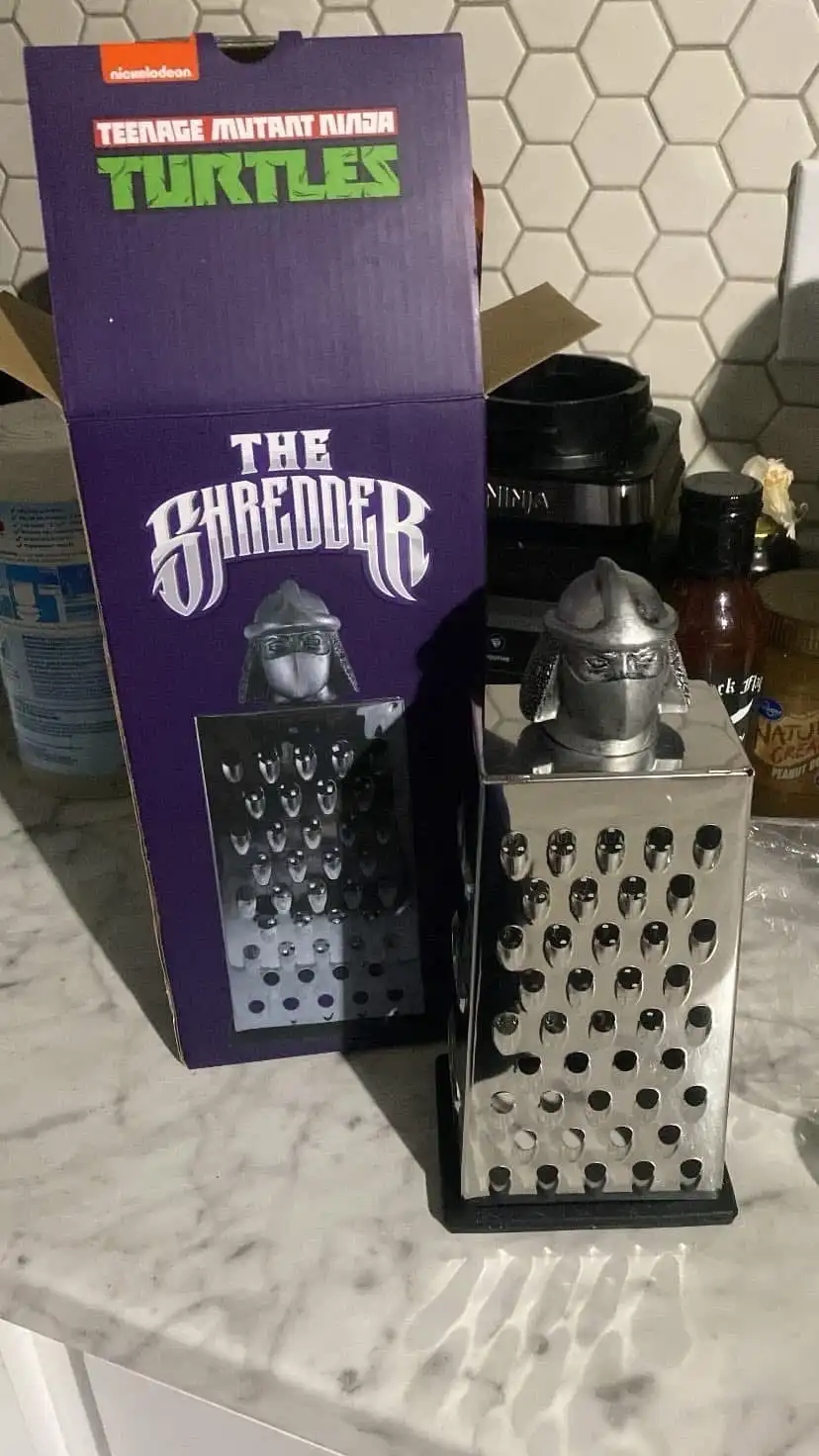 A metal box grater with a ninja helmet on top sits next to its original Teenage Mutant Ninja Turtles packaging, which…
