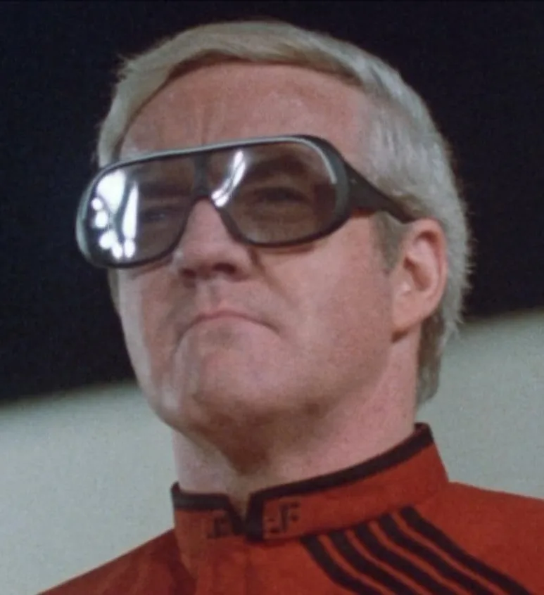 A man with gray hair wears large, thick-rimmed, futuristic-looking glasses while looking slightly upward with a skeptical or…