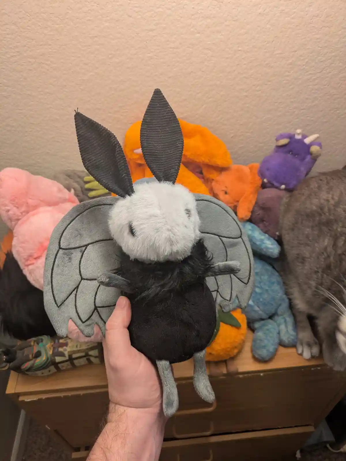A hand holds a plush toy resembling a bat with grey wings, black body, and long black ears, facing away from the camera…