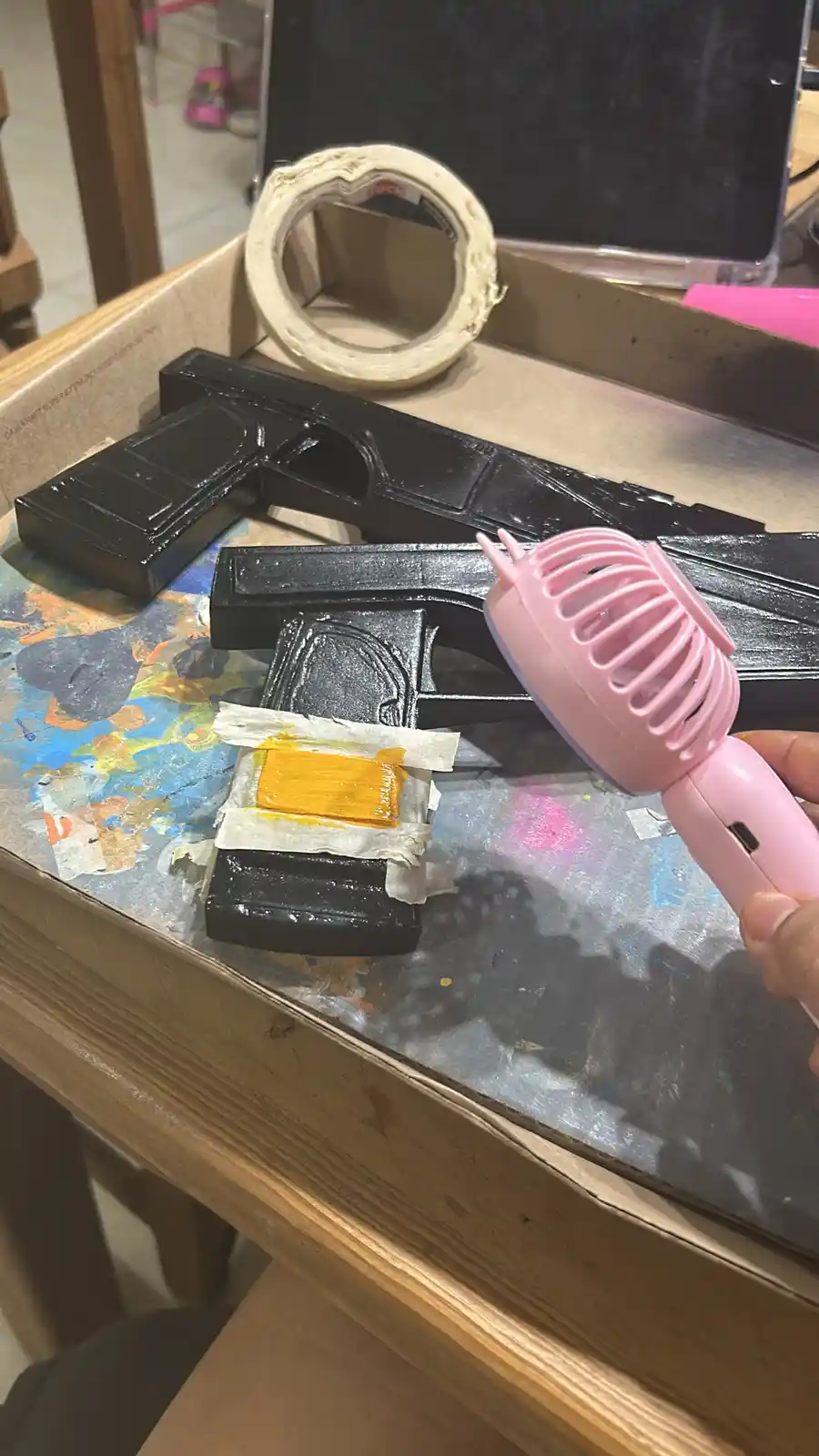 A hand holds a pink handheld fan over a black plastic gun-shaped object that has a rectangular patch of yellow paint taped…