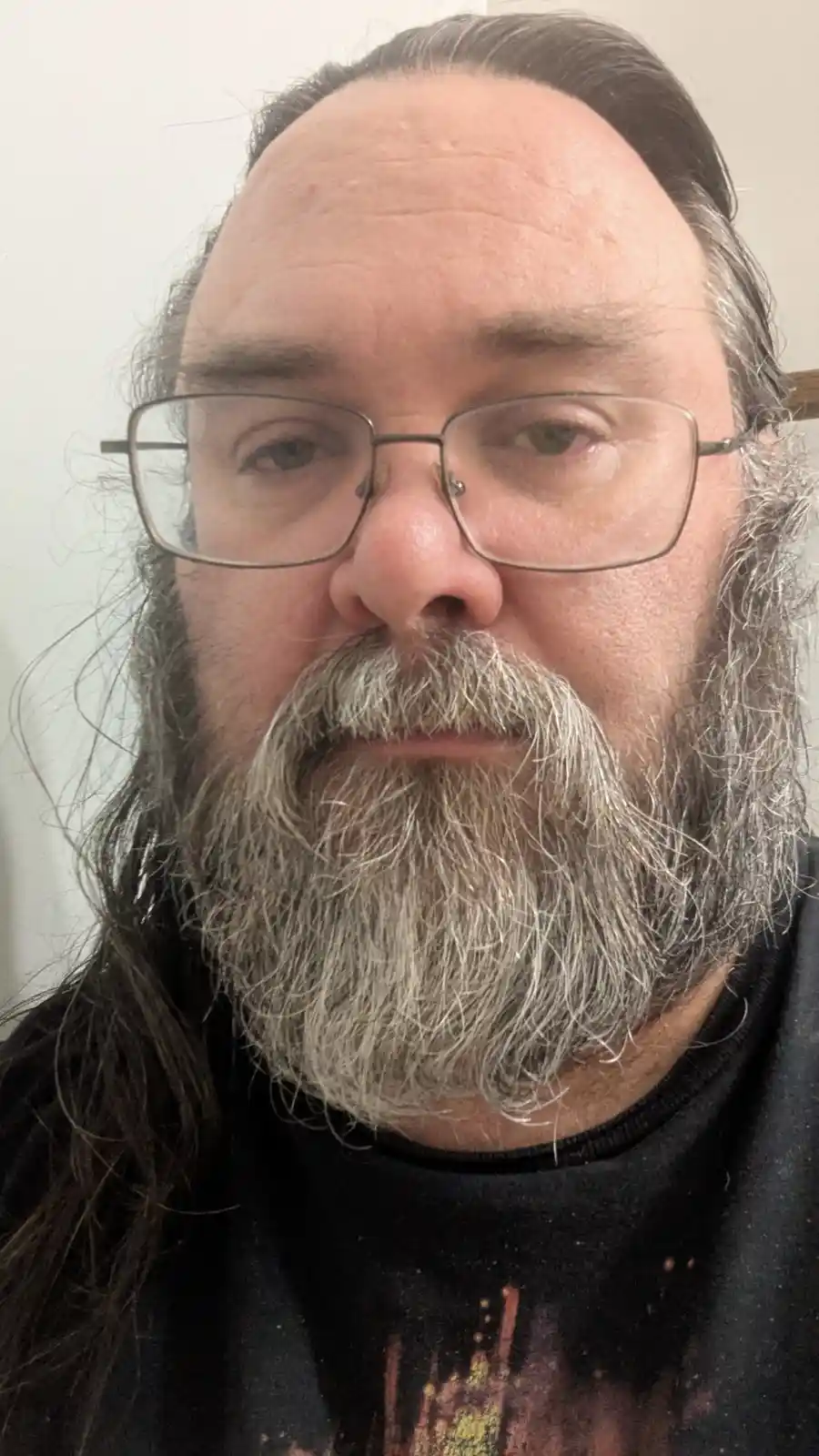A close-up photo of a middle-aged man with long dark hair, a full gray beard and mustache, and rectangular glasses. He is…