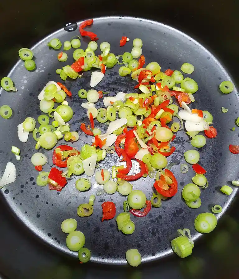 A close-up photo shows chopped green onions, red bell peppers, and garlic cloves scattered across the dark, speckled surface…