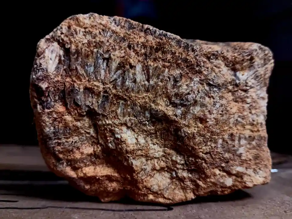a close-up photograph of a rough, brownish rock with a prominent cluster of translucent, needle-like crystals protruding…